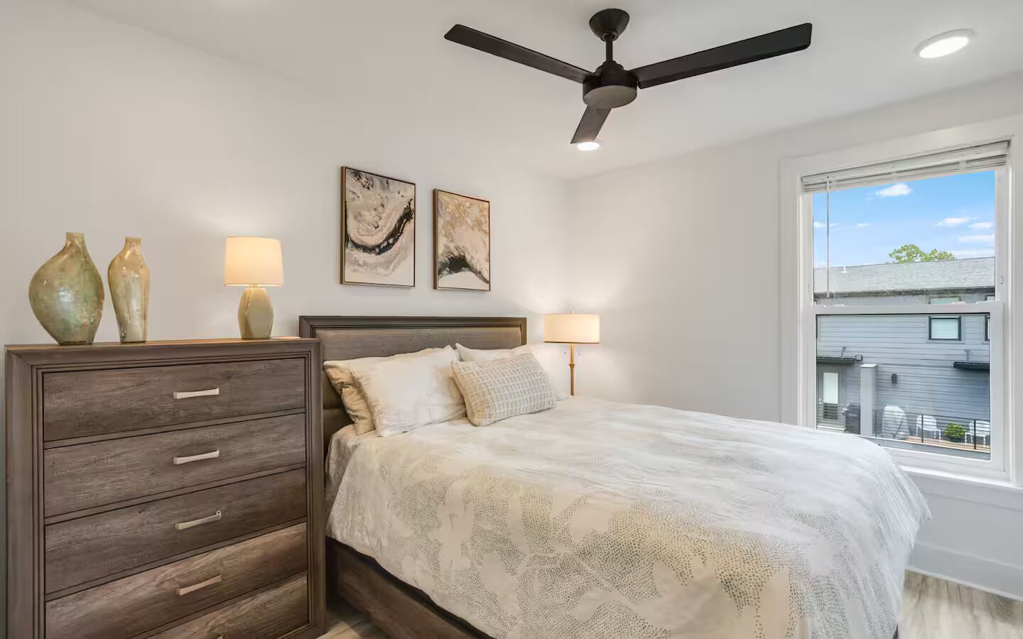 A bedroom with a bed, a dresser, two wall-mounted lamps, ceiling fan, and a window with a view.