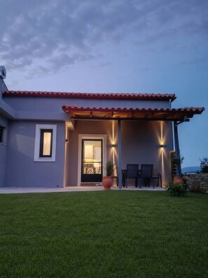 A modern house with a red-tiled roof, a well-lit entrance, and a small patio area with chairs and a table.