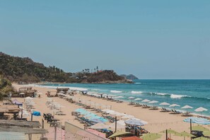 Ein Strand mit Reihen von Liegestühlen und Sonnenschirmen, ein klarer Blick auf den Ozean und eine Hügelkette mit Gebäuden im Hintergrund.