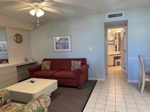 A living room with a red sofa, a white coffee table, a ceiling fan, a framed picture on the wall, and a kitchen visible through an open doorway. - Beachfront Condo at Treasure Island Beach Club (Treasure Island)