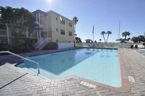 Ein Schwimmbecken mit Backsteinbord, ein Gebäude mit Balkon und Palmen im Hintergrund.