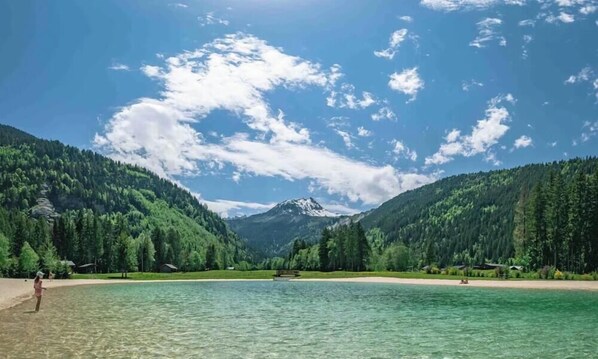 Ein See, umgeben von bewaldeten Bergen, unter wolkenlosem Himmel, mit einem entfernten Berggipfel.