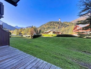 Eine Holzterrasse, eine grüne Rasenfläche und ein Gebäude mit rotem Dach in einer gebirgigen Gegend.