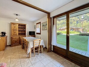 Ein Essbereich mit Tisch und Stühlen, einem Fernseher, Holzschränken und Blick auf eine grüne Landschaft durch eine Glastür.