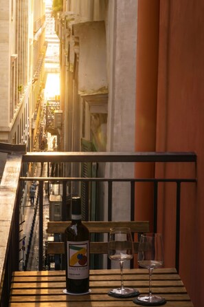 Un balcón con una botella de vino y dos copas sobre una mesa de madera, con vista a una calle estrecha flanqueada por edificios.