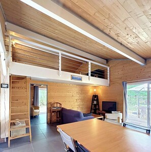 Ein Loft-Zimmer mit Holzbalkendecke, einem Esstisch, einer Couch, einem Fernseher und einem Fenster mit Blick ins Freie.