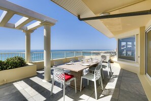 Um terraço com mesa e cadeiras, vista para o mar e um edifício ao fundo.