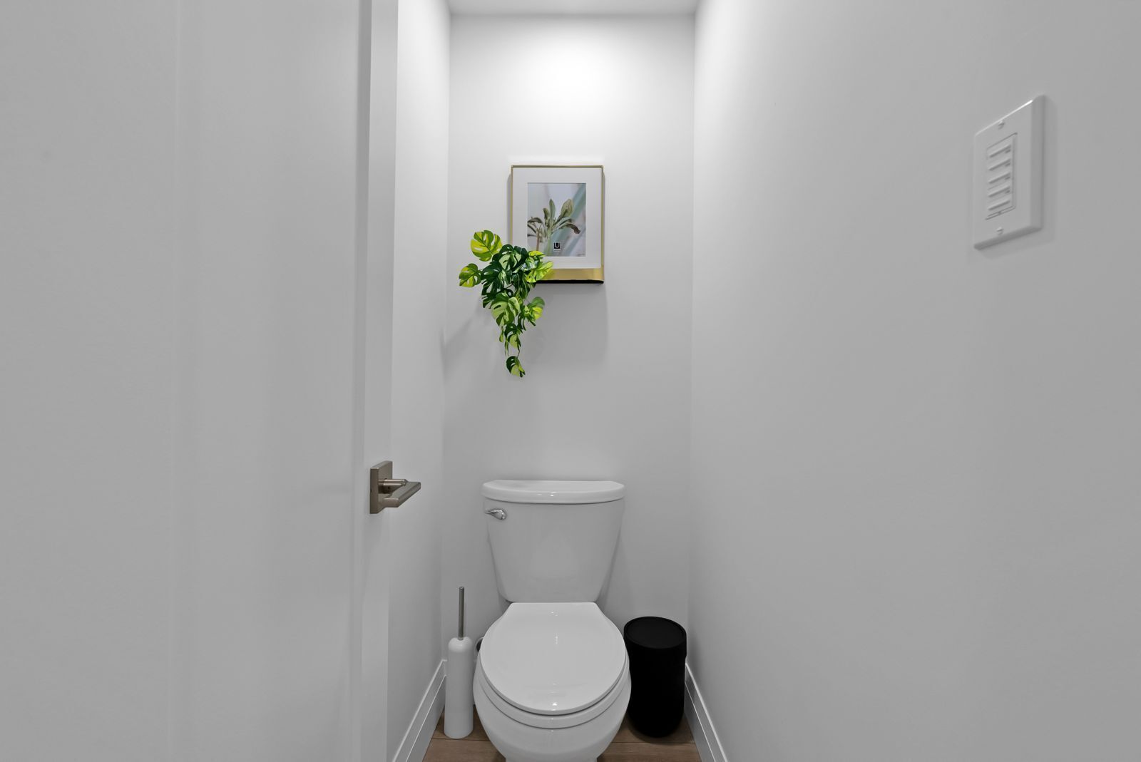 A small, white bathroom with a toilet, a trash bin, a toilet brush, and a framed picture on the wall.