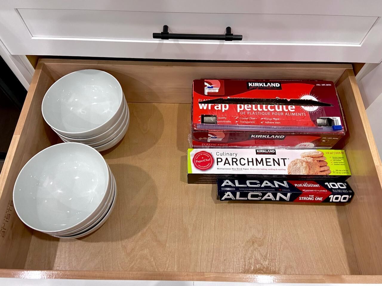 Eine Schublade mit weißen Schüsseln, einer Packung Kirkland-Folie, einer Packung Backpapier und zwei Packungen ALCAN-Papier.