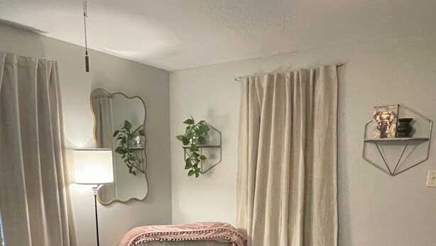 Quarto com cama, luminária de pé, ventilador de teto, mesinha de cabeceira, espelho e prateleira na parede.