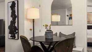 Ein Hotelzimmer mit einem runden Esstisch, zwei Stühlen, einer Lampe, einer Vase mit Blumen, einem großen Spiegel und einem schwarzen, abstrakten Kunstwerk an der Wand.