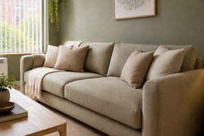 A beige sofa with pillows, a small table with a bowl and book, a potted plant, and a framed picture on the wall. - Marina Breeze Retreat 6 sleeps (Marina del Rey)