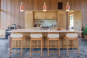 A modern kitchen with wooden cabinets, a central island with stools, pendant lighting, and a view of the outdoors. - Elsie's Hut: Mazama Cabin with Big Mountain Views (Mazama)