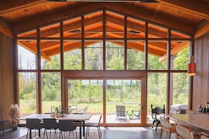 A spacious dining area with a large table and chairs, large windows offering a view of the surrounding forest, and a high wooden ceiling. - Elsie's Hut: Mazama Cabin with Big Mountain Views (Mazama)