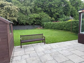 A wooden bench on a paved patio with a grassy lawn and a wooden fence in the background.