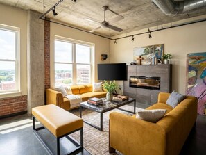 A modern living room with a sofa, armchair, and ottoman, a flat-screen TV, and a fireplace. - Industrial Loft · Walk to Westport · Sleeps 6 (Kansas City)