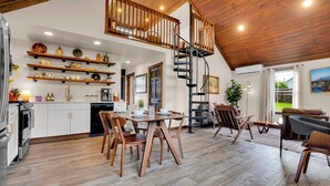 Une cuisine spacieuse avec des meubles en bois, un coin repas avec une table et des chaises en bois, un escalier en colimaçon et une vue sur l’extérieur.