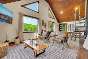 A spacious living area with a wooden ceiling, large windows, a staircase, and a view of greenery.