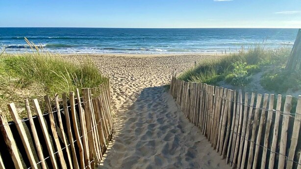 Ein sandiger Küstenweg führt zum Meer, begrenzt von Holzzaun.