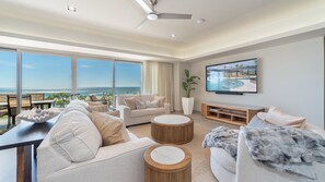 Ein modernes Wohnzimmer mit großem Fenster mit Blick auf einen Strand, einem Deckenventilator, einem Flachbildfernseher und einer Holz-Unterhaltungsanlage.