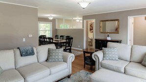 A living room with a beige sofa set, a dining table with chairs, a wooden coffee table, and a mirror on the wall.