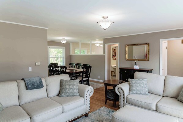 A living room with a beige sofa set, a dining table with chairs, a wooden coffee table, and a mirror on the wall.