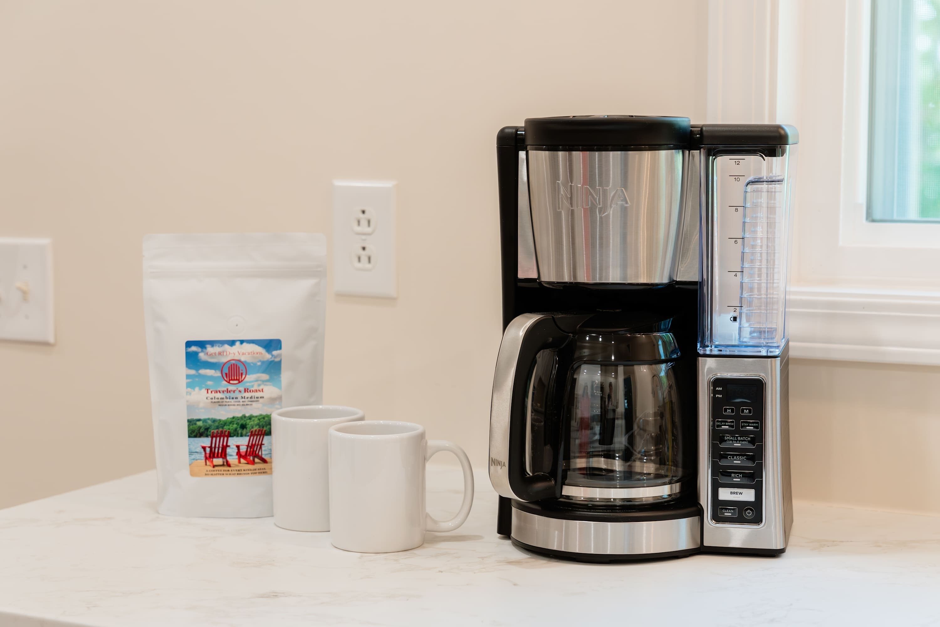 Máquina de café com jarra e bandeja coletora, um saco de café e duas canecas brancas em uma bancada de cozinha.