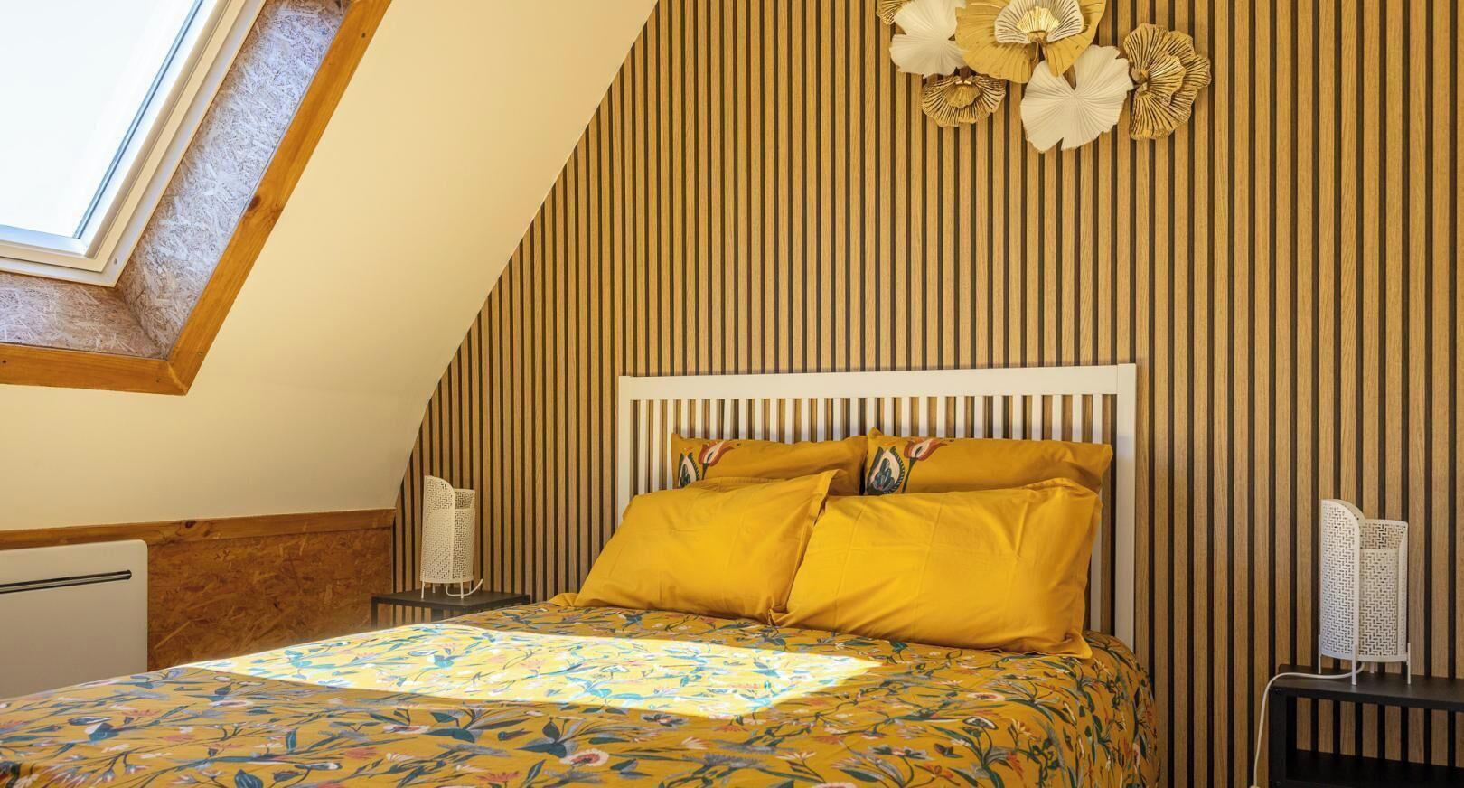 Um quarto com teto inclinado, uma cama com colcha florida, cabeceira branca e parede com ripas de madeira verticais.