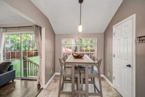 A dining area with a table and chairs, a hanging light fixture, a sliding glass door leading to a backyard, and a white door. - Island Home. 15 mins to Tybee Beach & Downtown., Sleeps 10 (Savannah)