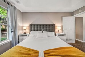 A bedroom with a large bed, two bedside tables, a window with curtains, and a door. - Island Home. 15 mins to Tybee Beach & Downtown., Sleeps 10 (Savannah)