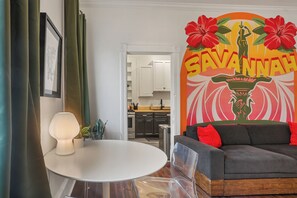 A living room with a white round table, a grey sofa with red cushions, a framed picture on the wall, and a decorative wall piece with the word "SAVANNAH". - Savannah Home, Walkable to Forsyth, Historic, Dogs OK, Free Parking for 2 Cars (Savannah)
