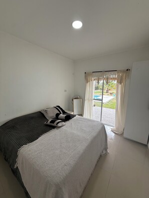 A bedroom with a bed, a bedside table, a fan, and a view of the outside.