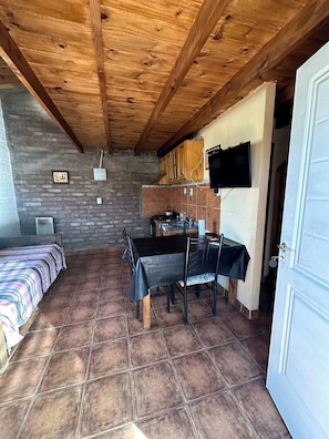 A room with a wooden ceiling, a dining table with chairs, a TV mounted on the wall, and a bed with a striped bedspread.