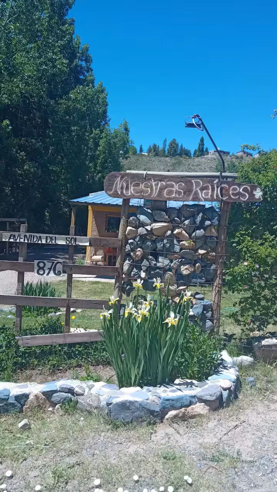A wooden sign with 'Nuestras Raices' written on it, surrounded by rocks and yellow flowers.