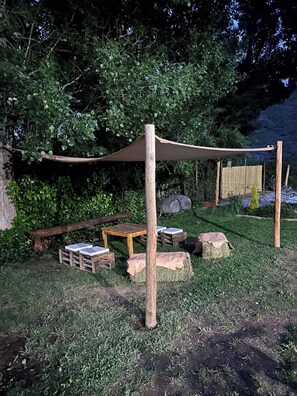 A rustic outdoor seating area with wooden tables and benches, surrounded by greenery and a shaded canopy.