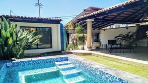 En swimmingpool med blå mosaikfliser, en terrasse med udendørs møbler og en bygning med et tag af fliser.
