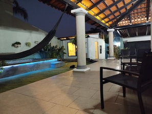 A covered outdoor area with a hammock, a pool with blue lighting, and a seating area with chairs.