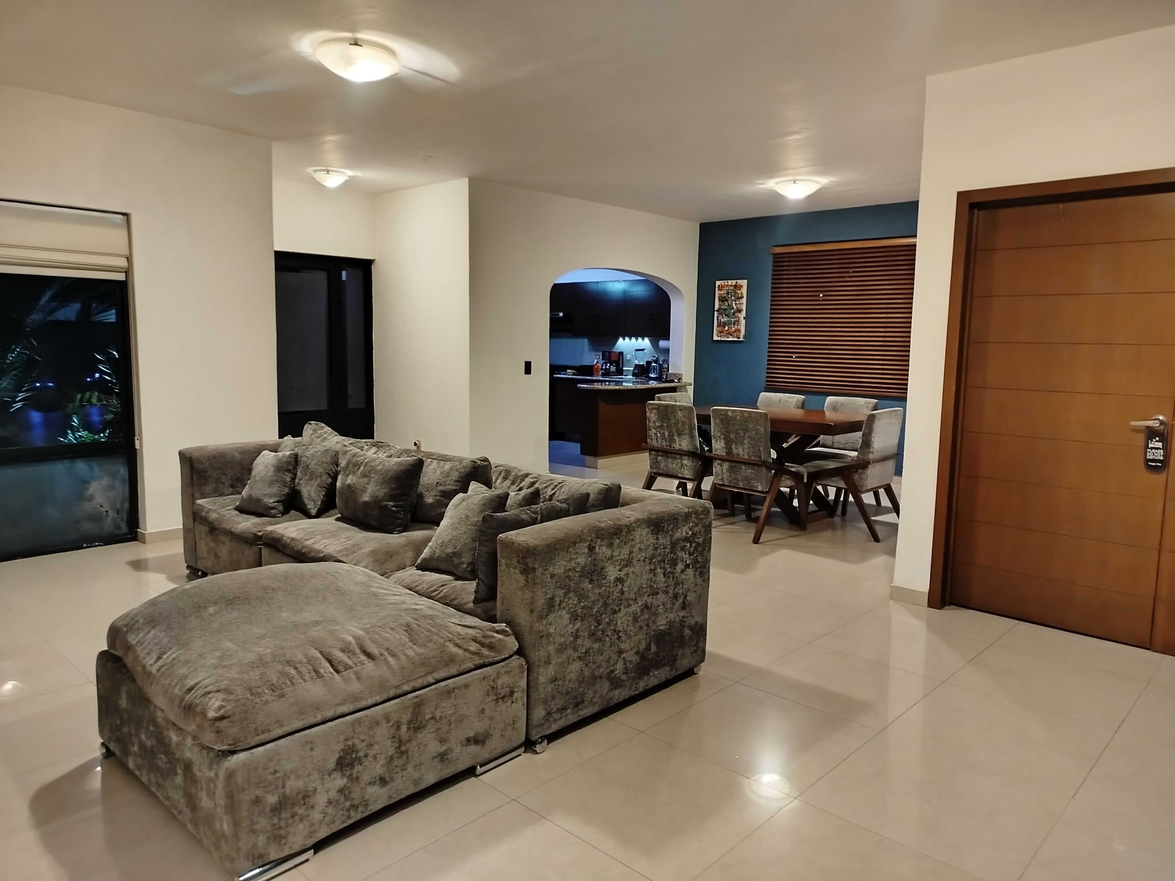 A living room with a sectional sofa, a dining area with a table and chairs, and a kitchen in the background.