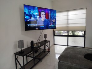 A living room with a flat-screen TV mounted on the wall, a black entertainment unit with speakers, and a large window providing natural light.