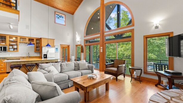 A spacious living area with a large sofa, a coffee table, and a TV. There is a kitchen area with wooden cabinets and a dining table. Large windows provide natural light and views of the outdoors. - Mi Casa, Su Casa | Fire Pit | Dock | Game Rom | Sleeps 14 (Sherrills Ford)