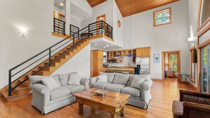 A spacious living area with a wooden staircase, a kitchen in the background, and a large window providing natural light. - Mi Casa, Su Casa | Fire Pit | Dock | Game Rom | Sleeps 14 (Sherrills Ford)