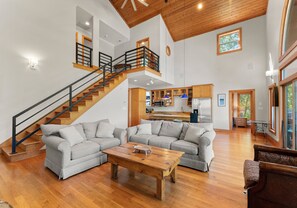 A spacious living area with a wooden staircase, a kitchen in the background, and a large window providing natural light. - Mi Casa, Su Casa | Fire Pit | Dock | Game Rom | Sleeps 14 (Sherrills Ford)