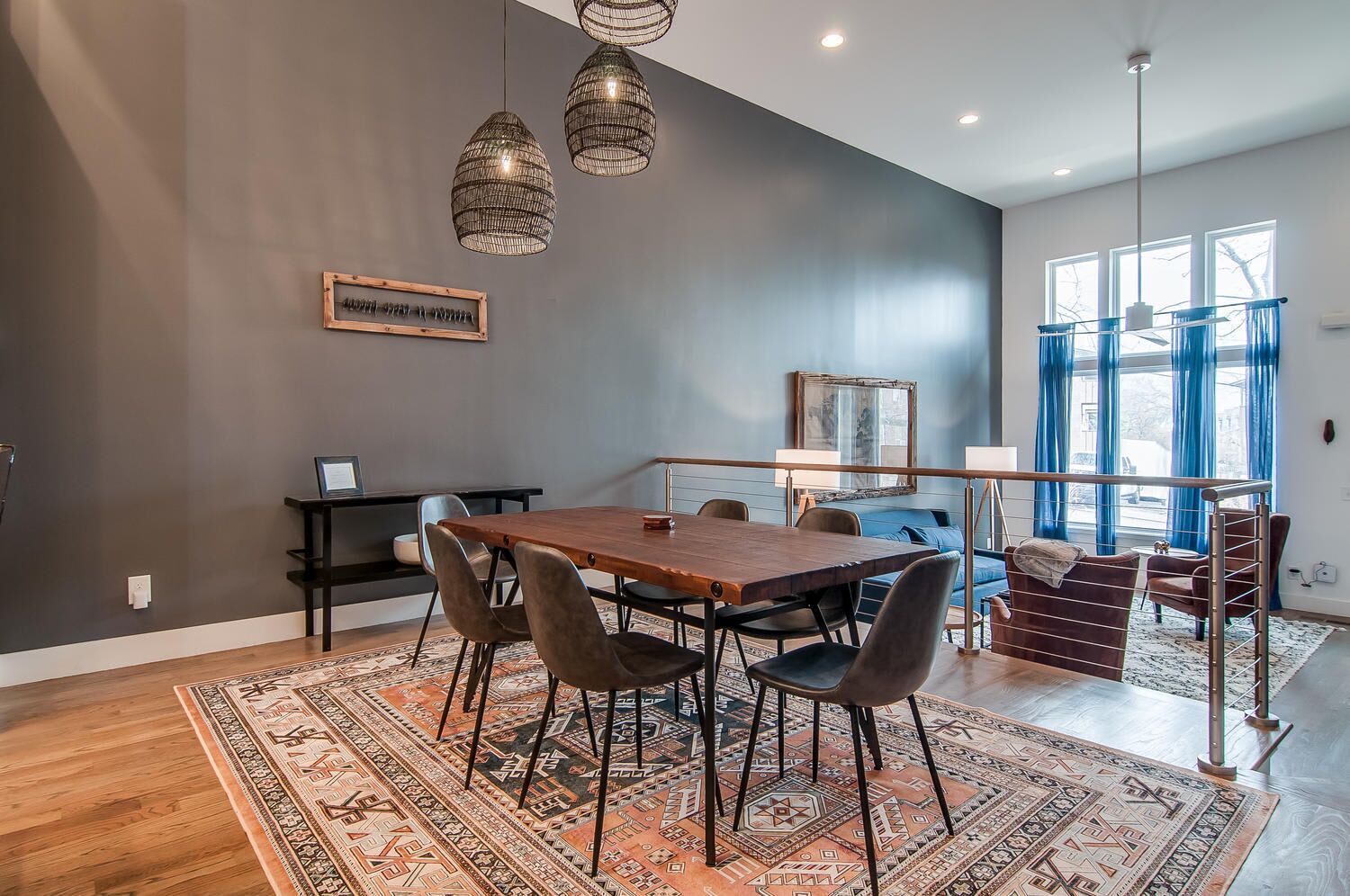 A modern dining area with a wooden table, chairs, a patterned rug, pendant lights, and a view of the outdoors through large windows.