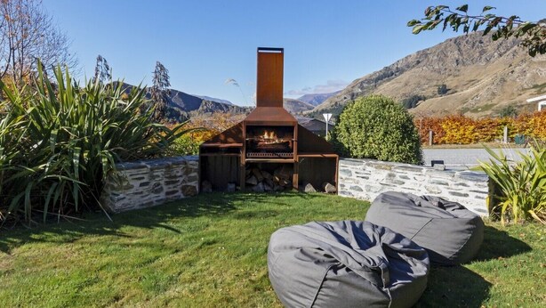 A stone wall, a wooden barbecue, and a grassy area with a view of mountains. - Perfect for families, this spacious home with spa pool is perfect for relaxation or adventure. (Queenstown)