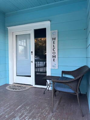 Eine Veranda mit einem 'WELCOME'-Schild, einem Korbstuhl und einer Tür mit Fenster.