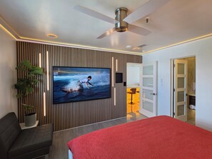 Et moderne hotelværelse med et stort fladskærms-tv, en ventilator i loftet, en seng med rødt sengetøj, et siddeområde med en stol og en potteplante.