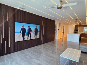 En moderne hotellobby med et stort tv, der viser et strandmotiv, en stilren receptionsdisk og et tidssvarende design med træpaneler og indbygget belysning.