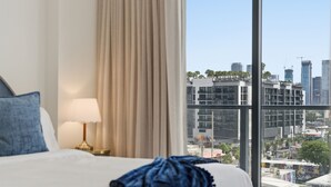 Habitación de hotel con cama, lámpara, cortina y un amplio ventanal con vista a la ciudad.