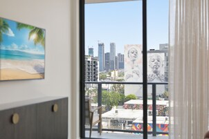 A room with a large window overlooking a cityscape, a painting of a beach, and a chair.