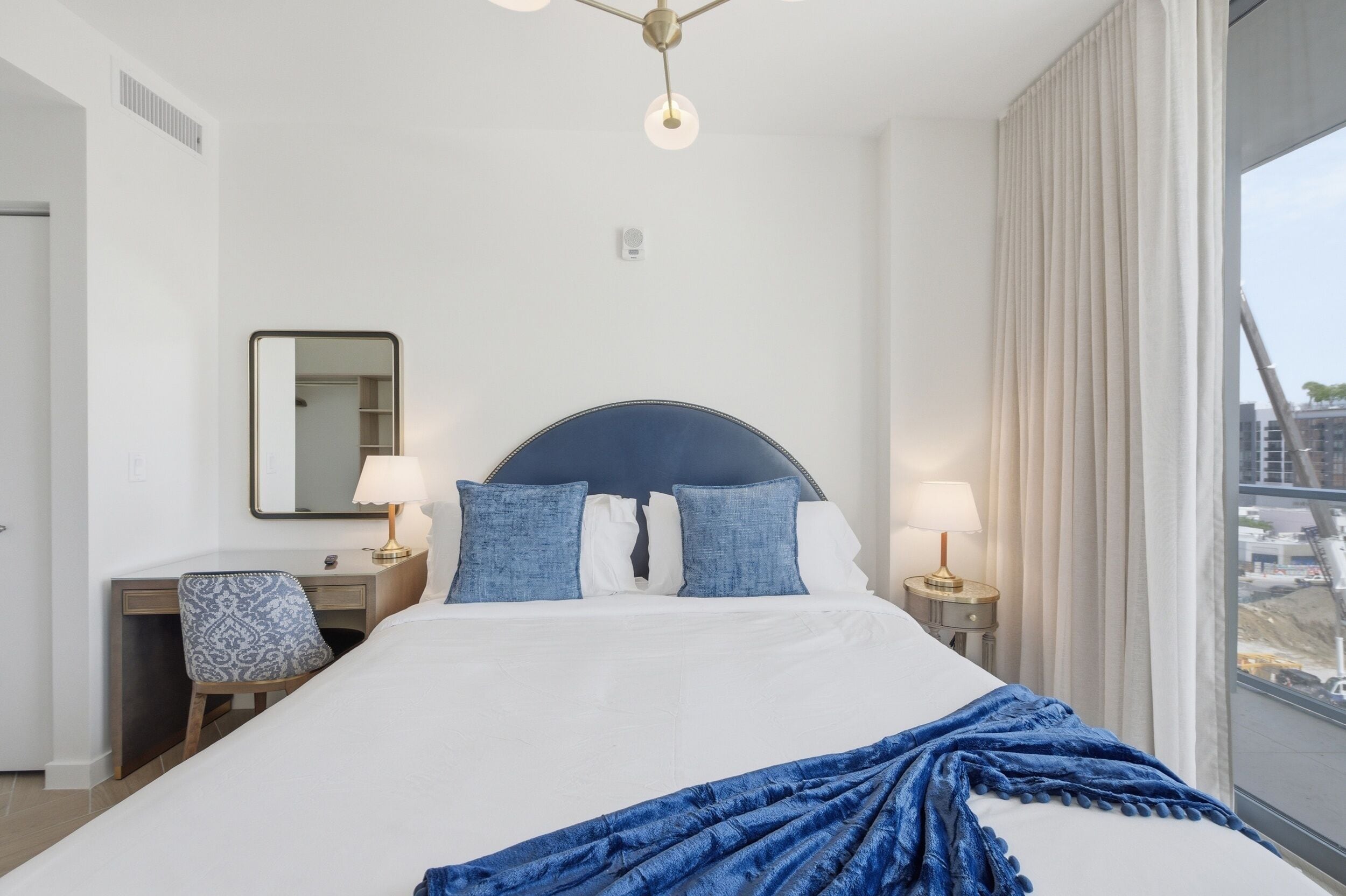 A hotel room with a bed, bedside lamps, a desk, a chair, a mirror, and a view of the outside.